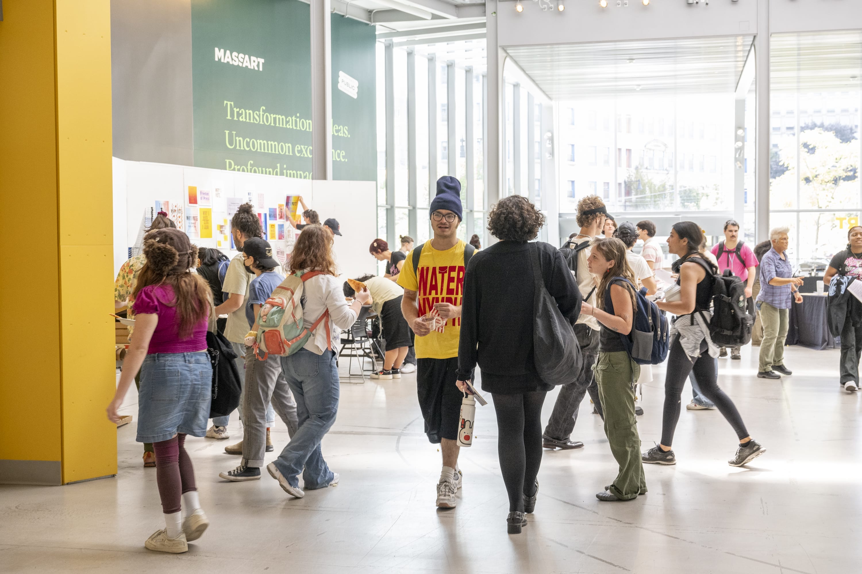 A busy, sunlit lobby at MassArt with students walking and talking. People wear casual clothes and backpacks. Colorful art is displayed on a white wall, and large windows let in natural light. A green MASSART sign is visible in the background.