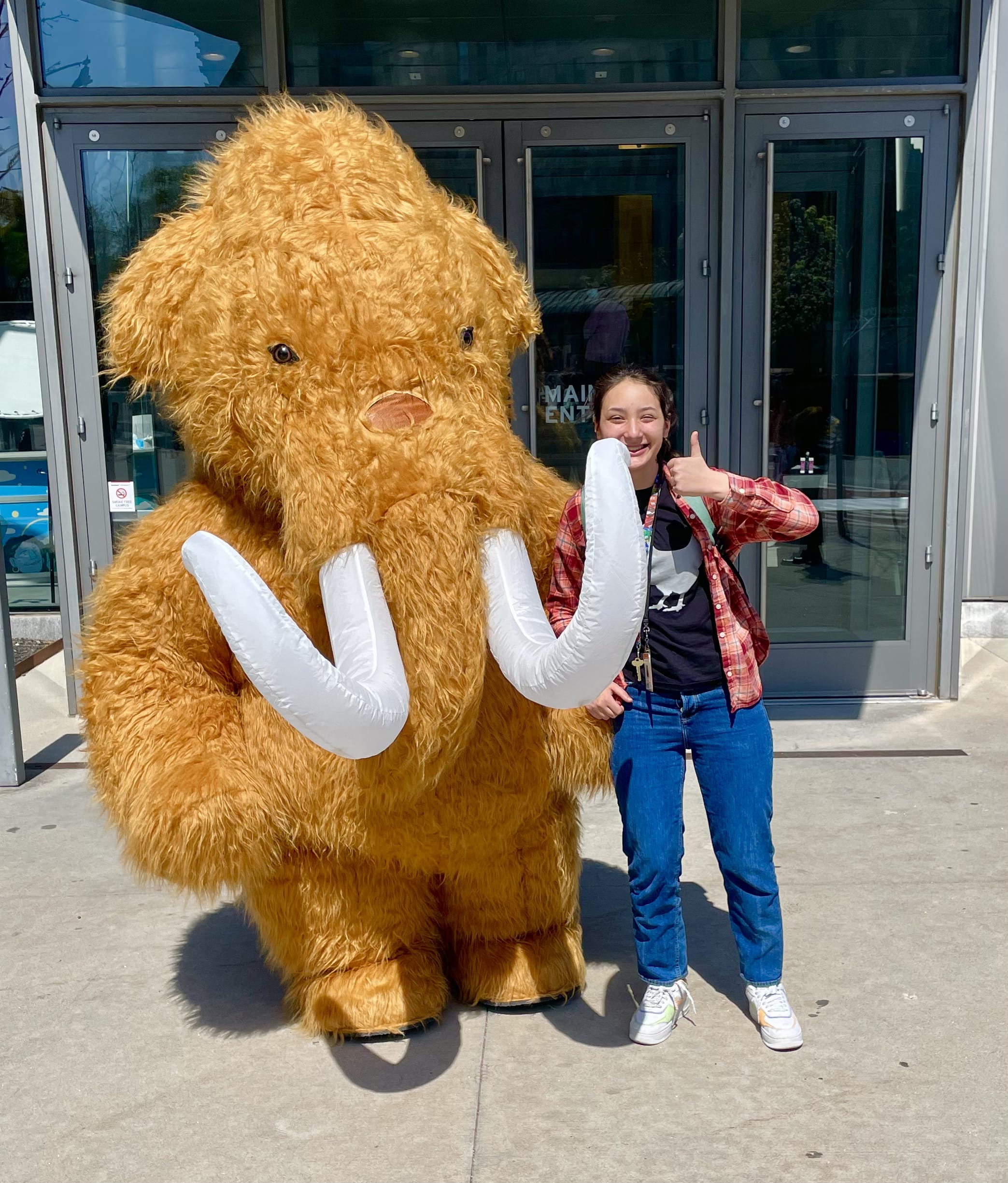 Student leader Kailey Caballero smiles and gives a thumbs-up while standing next to someone in a large, fuzzy woolly mammoth costume with big white tusks, outside a building with glass doors on a sunny day.