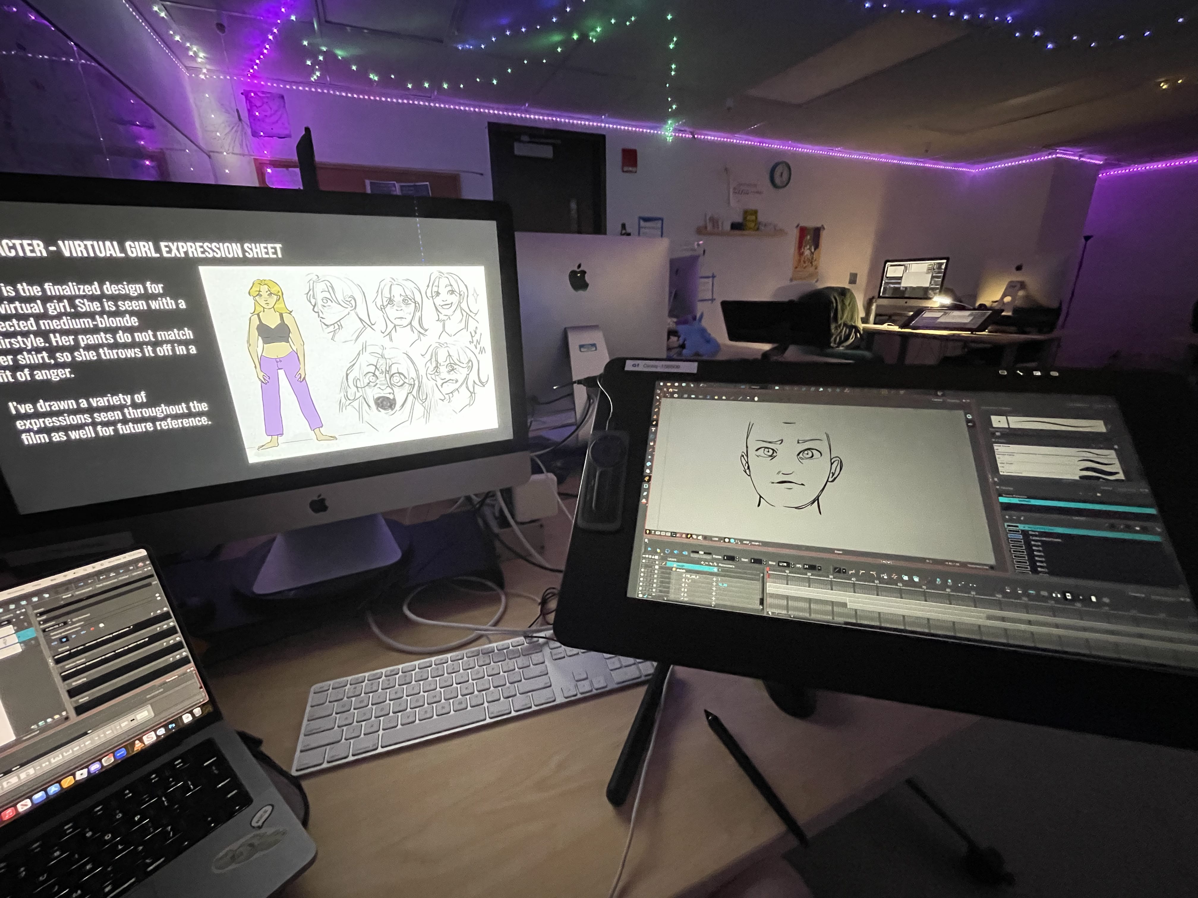A dimly lit workspace with multiple monitors displays digital art: one features student leader Kailey Caballero’s animation character design sheet of a girl, while another shows a drawing in progress. Colorful string lights decorate the ceiling.