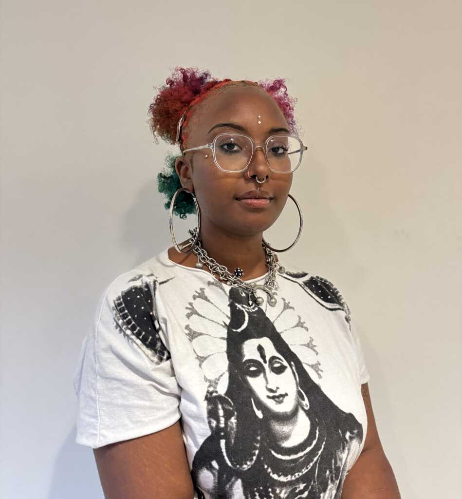 A person with colorful hair, glasses, nose and septum rings, large hoop earrings, and a chain necklace wears a white t-shirt featuring an illustration of a deity, standing against a plain light background at the MassArt Board of Trustees event.