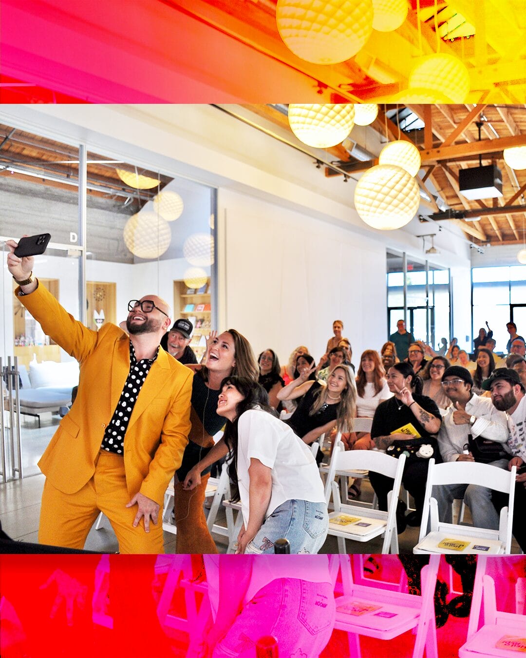 MassArt alum Joseph Soto, in a bright yellow suit, takes a group selfie with an enthusiastic audience seated on white chairs in a modern event space with hanging lantern lights and wooden ceilings.
