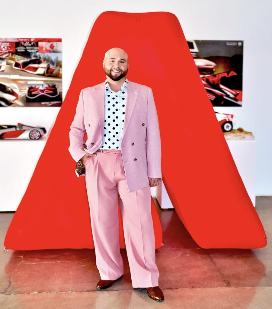 MassArt alum Joseph Soto, a smiling man in a pink suit, light blue polka dot shirt, and brown shoes, stands before a large red geometric sculpture and car design posters in a gallery setting.
