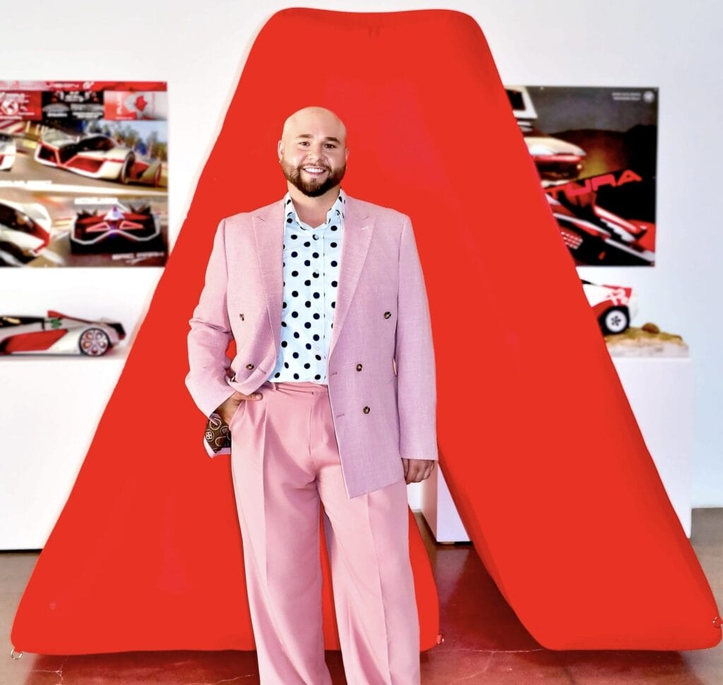 MassArt alum Joseph Soto, a smiling man in a pink suit, light blue polka dot shirt, and brown shoes, stands before a large red geometric sculpture and car design posters in a gallery setting.