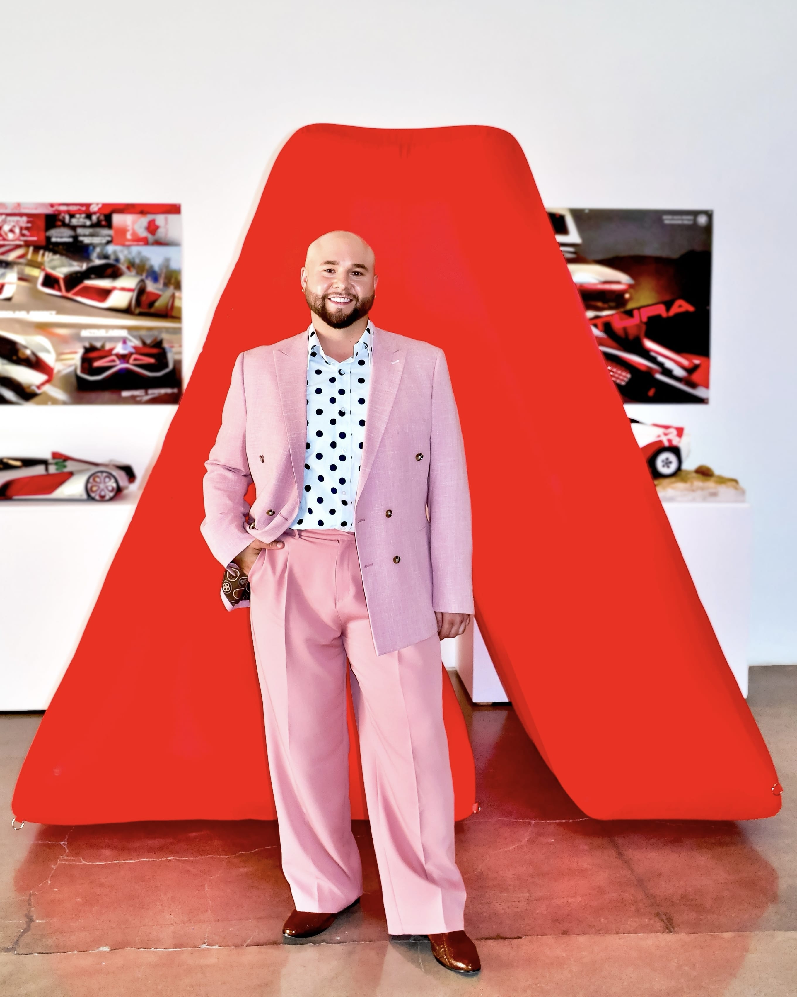 MassArt alum Joseph Soto, a smiling man in a pink suit, light blue polka dot shirt, and brown shoes, stands before a large red geometric sculpture and car design posters in a gallery setting.