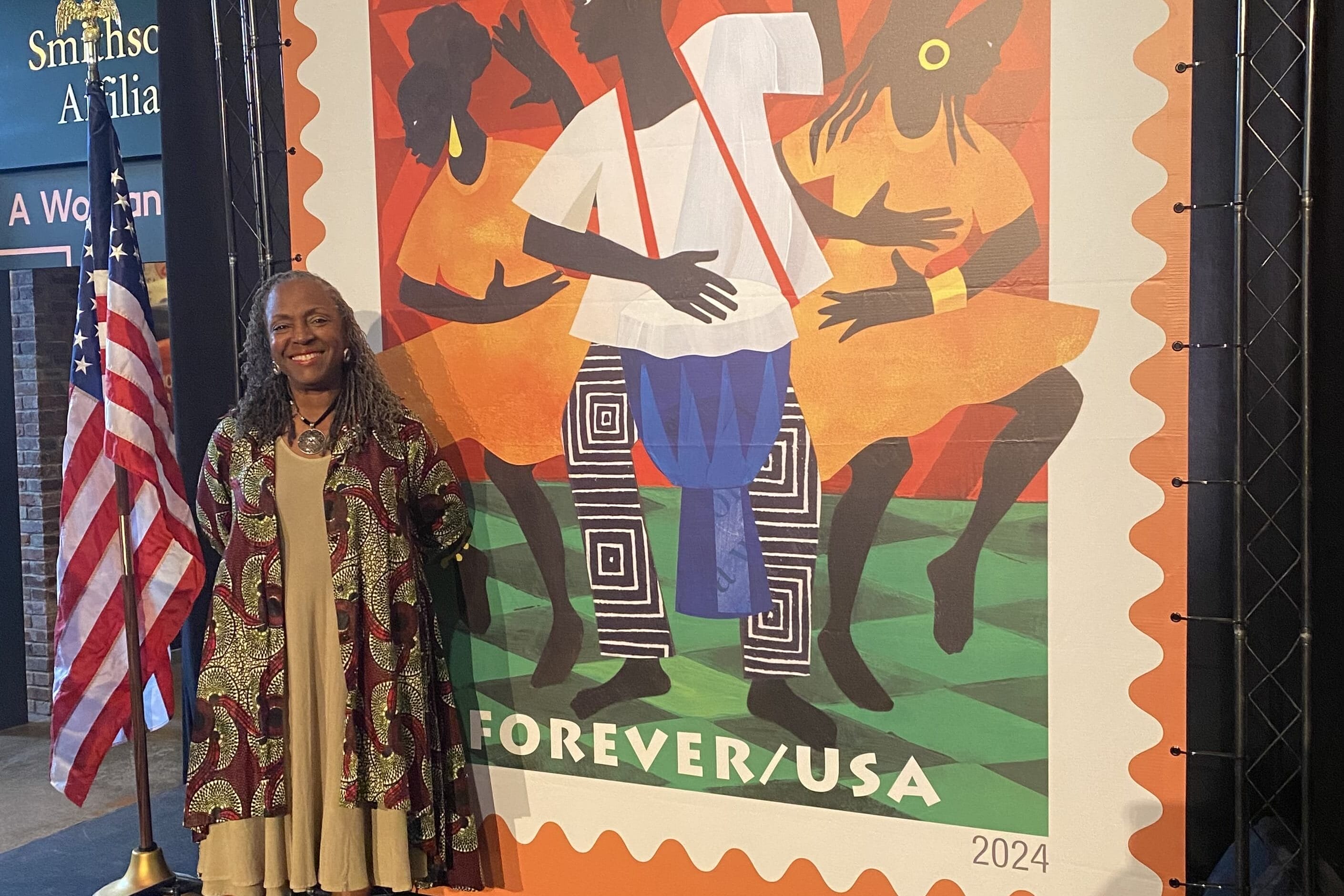 A woman smiles beside a vibrant Kwanzaa 2024 Forever USA postage stamp designed by Ekua Holmes, featuring stylized figures dancing and playing drums. An American flag and Smithsonian signage appear in the background, celebrating the lasting impact of culture.