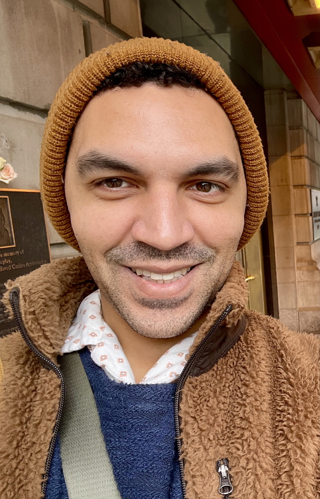 A person wearing a brown knit beanie, brown fleece jacket, and patterned shirt smiles at the camera outside near a brick building.