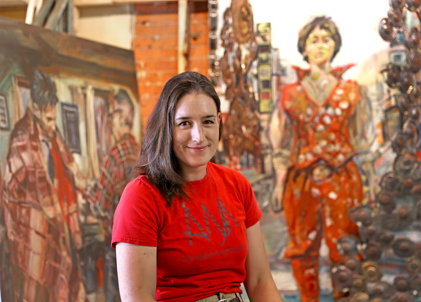 A MassArt alumna in a red t-shirt sits and smiles in an art studio, surrounded by large, colorful paintings and exposed brick walls in the background.