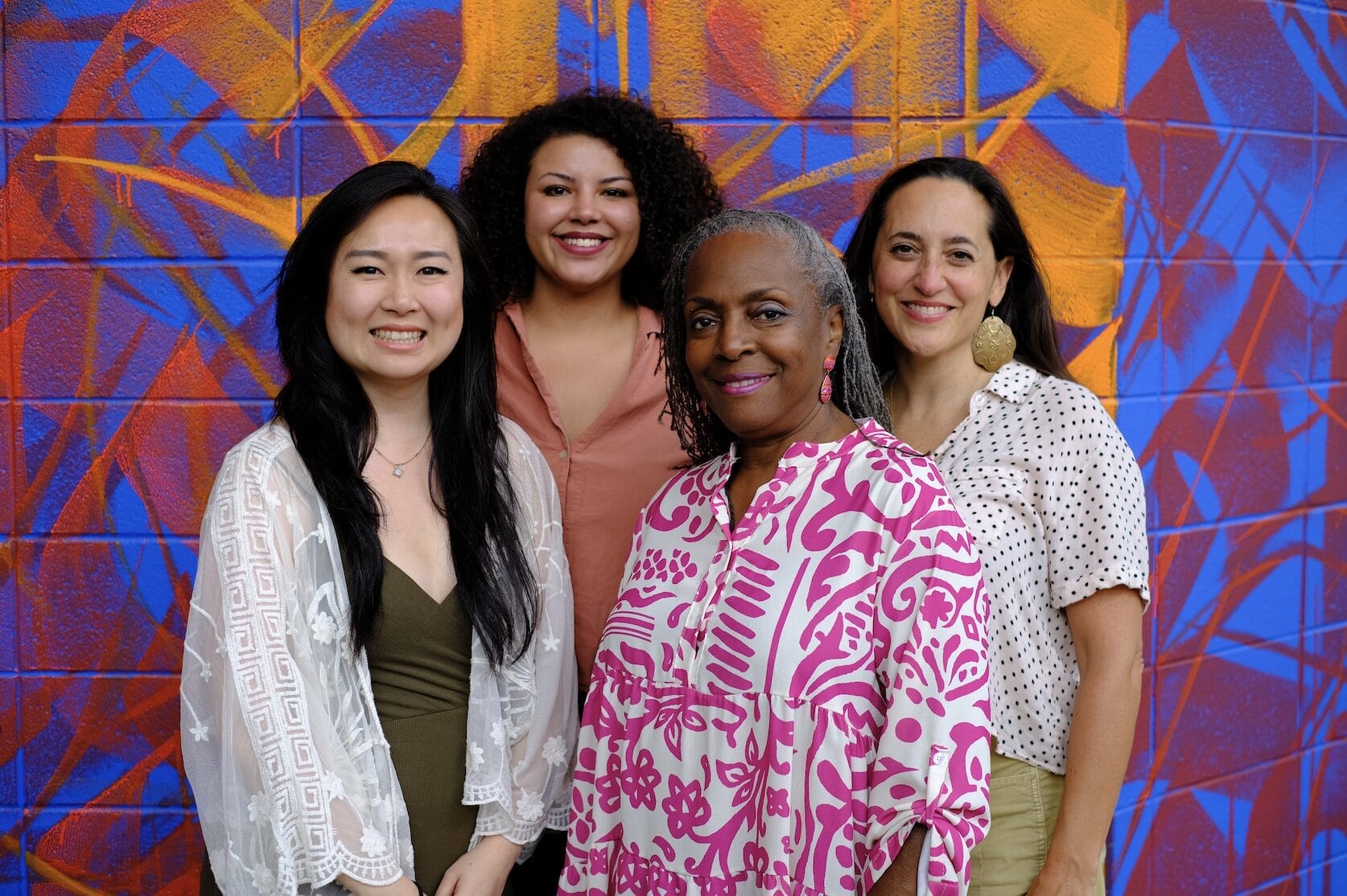 Four women stand together smiling in front of a colorful mural at the art center, dressed in casual, patterned clothing. Their happiness and relaxed vibe reflect the spirit of community partnerships.