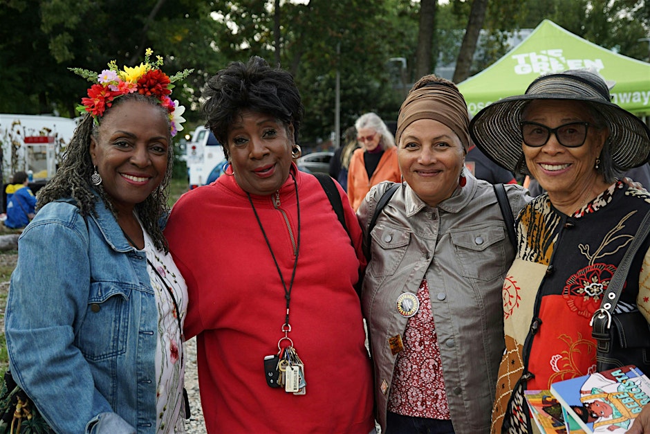 Four smiling women pose together outdoors at a MassArt event. One wears a flower crown, another a red top, a third a headscarf, and the last large sunglasses and a wide hat—a scene with Ekua Holmes-style warmth that feels like a miracle.