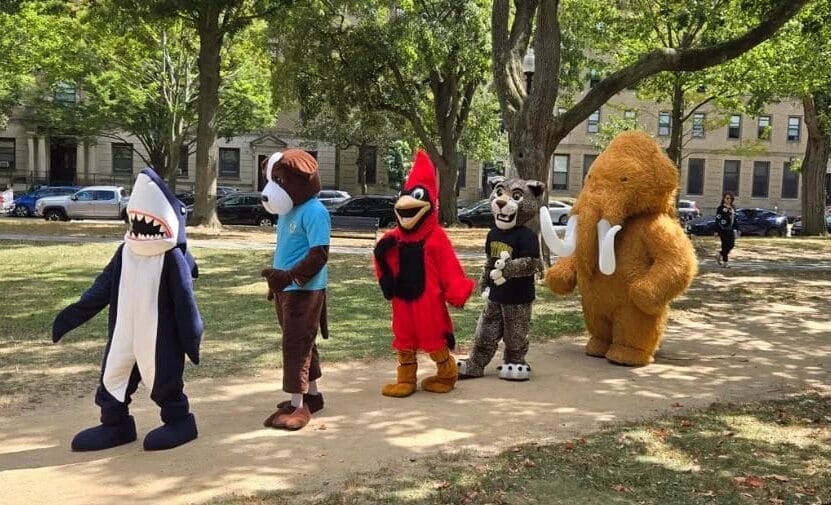 Five mascots—a shark, a dog, a red bird, a wildcat, and a furry mammoth—walk in a line outside on a tree-lined path at the Colleges of the Fenway, with buildings and parked cars in the background.