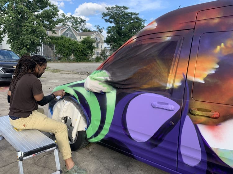 A person sits on a bench, painting colorful abstract designs in purple, green, and orange on sparc! the ArtMobile van parked outdoors, with houses and trees in the background.