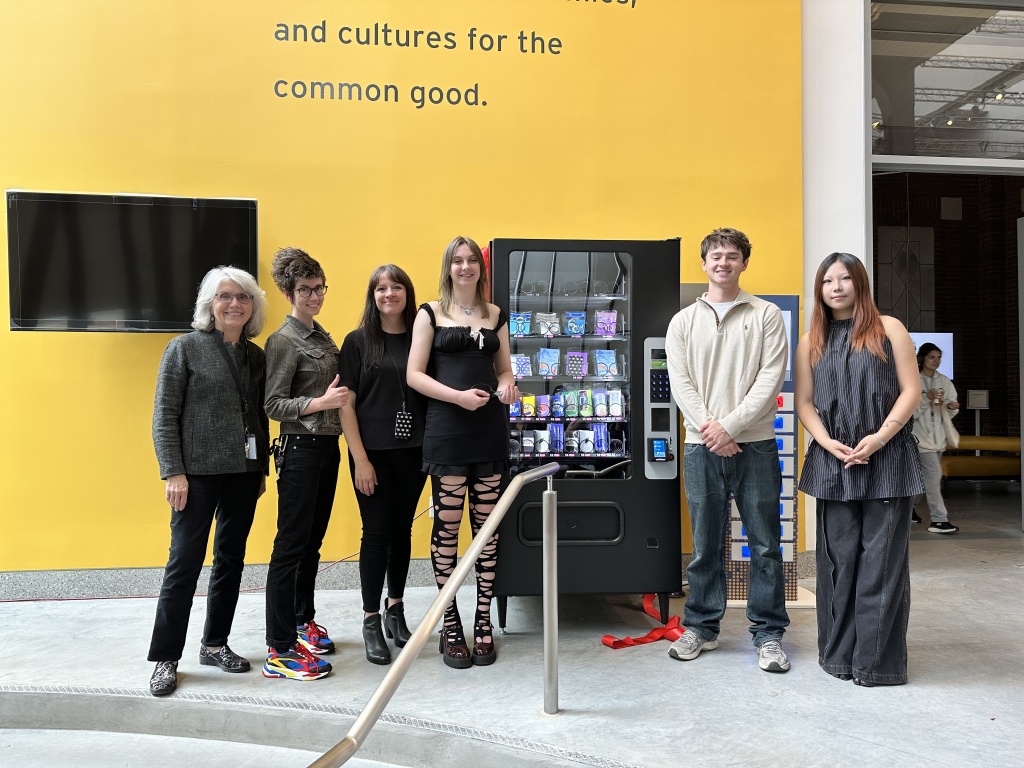 Six people stand in front of a vending machine and a bright yellow wall with a partial quote about the common good. Featured in the President's Newsletter, they are posing and smiling, with a velvet rope barrier in front of them.