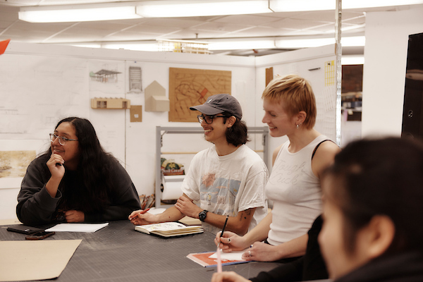 Four people sit and smile around a table in a bright, casual workspace, with notebooks and papers in front of them, suggesting a collaborative Admissions Meeting or discussion.