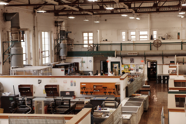 A large industrial workshop with high ceilings, workstations, printmaking equipment, and ventilation pipes hosts Admissions Meetings as people work at desks; natural light streams in from tall windows.
