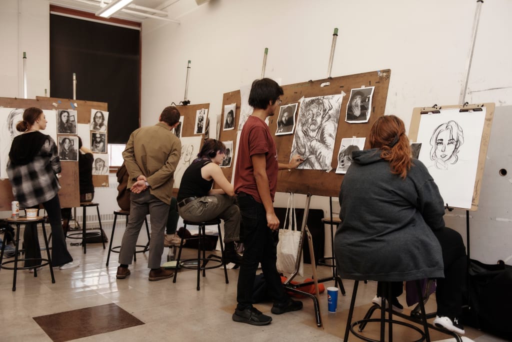 Several people sit and stand at easels in an art classroom, drawing black-and-white portraits on large sheets of paper. Finished and in-progress sketches are visible as the group prepares for upcoming Admissions events.