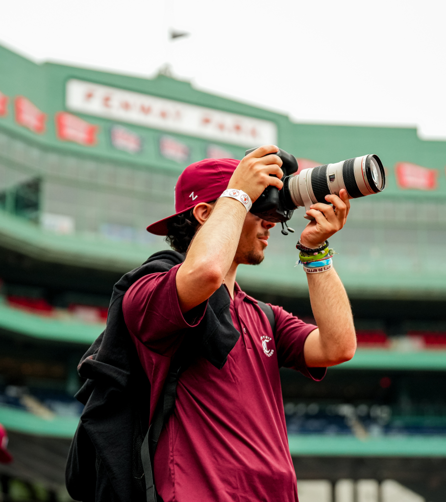 A student photographer in a maroon shirt and cap is taking a photo with a large camera at Fenway Park, with the stadium’s green stands and blurred signage visible in the background.