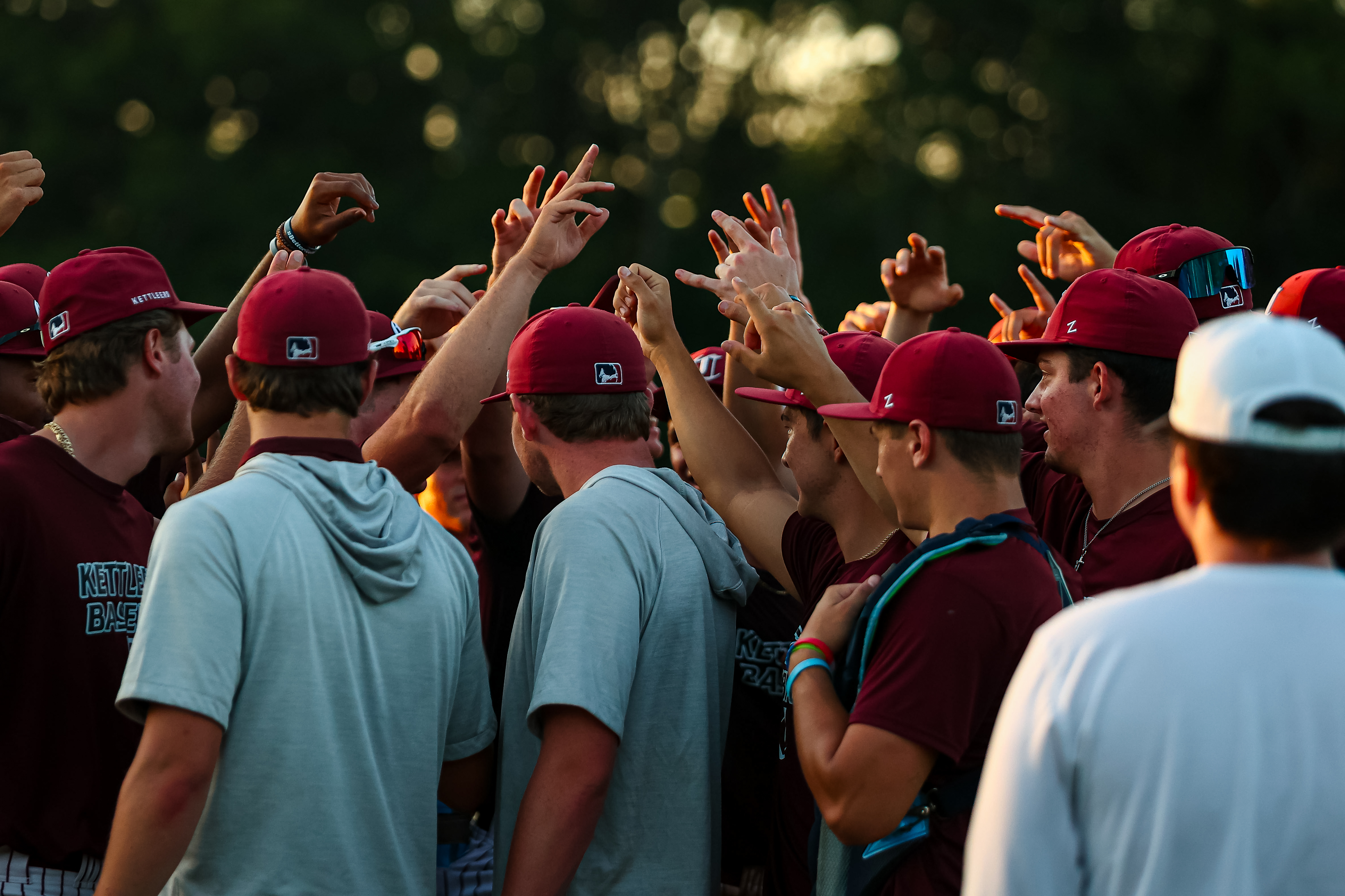 A group of Cape Cod Baseball players wearing maroon hats and shirts huddle together, raising their hands in unity. Captured behind the lens by a MassArt student, trees blur softly in the background.