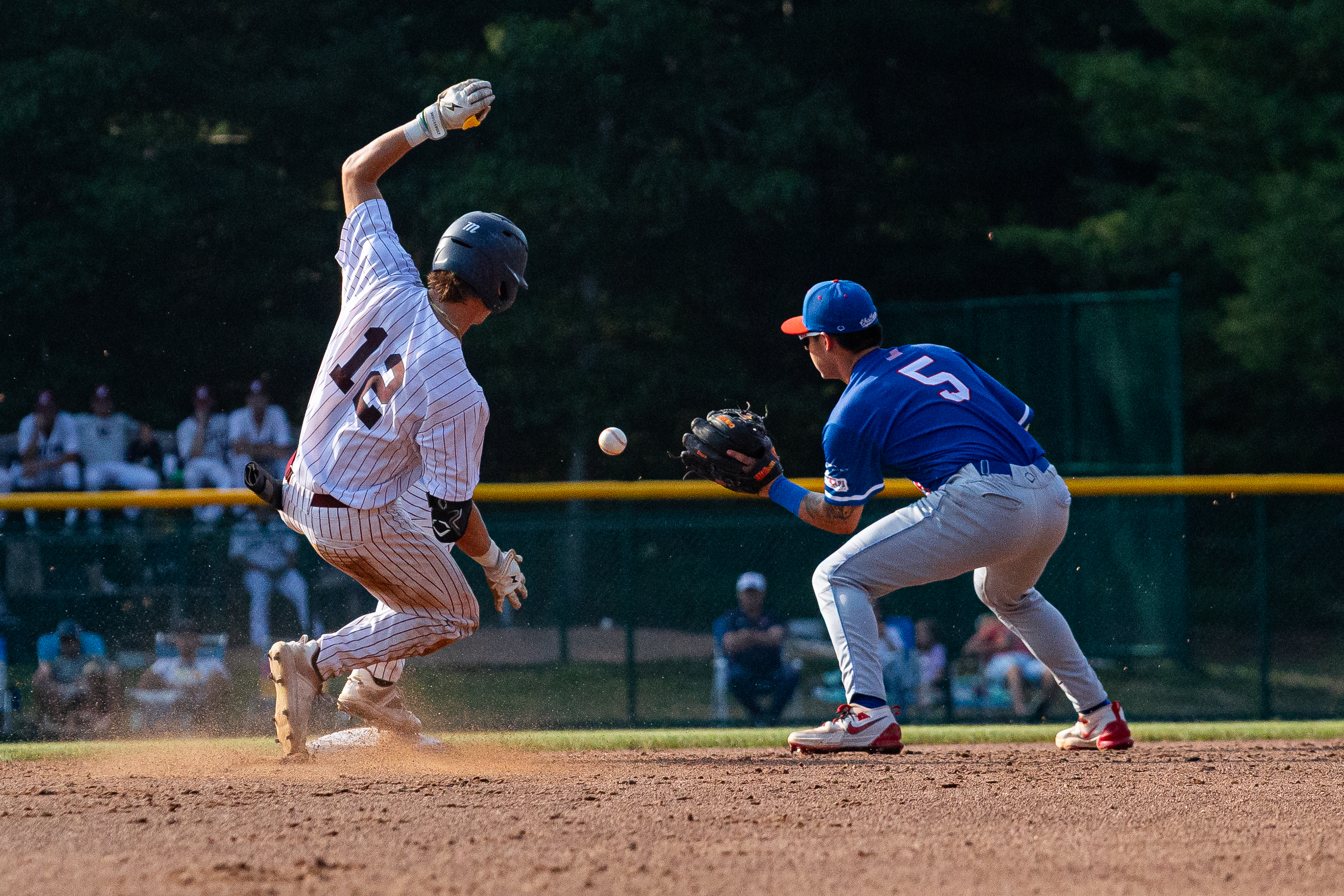 A Cape Cod Baseball player in a white pinstripe uniform slides toward a base as a player in blue waits to catch the ball. Behind the Lens, blurred spectators and green trees complete the action-filled scene.