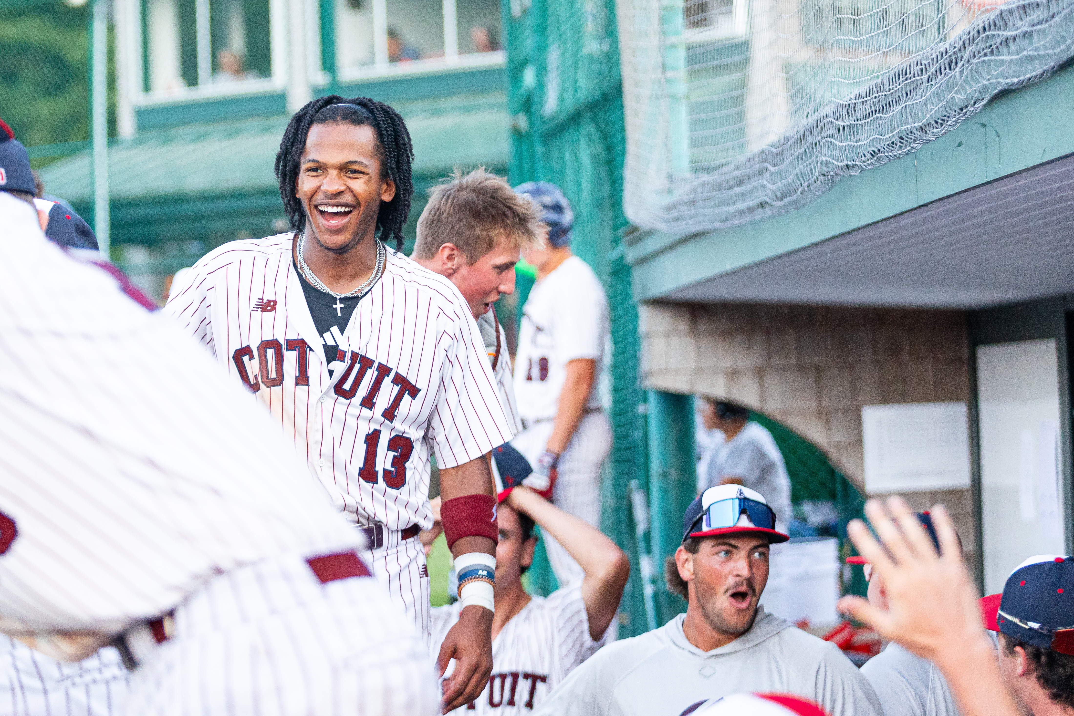 A smiling baseball player in a white-striped Cotuit uniform stands near the dugout, surrounded by cheering teammates, celebrating during a Cape Cod Baseball game captured Behind the Lens by a MassArt Student.