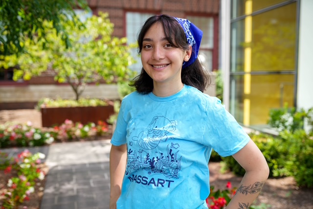 A young person with dark hair and a blue bandana smiles outdoors, wearing a light blue MassArt t-shirt with an illustrated design, standing on a path near flowers and greenery.