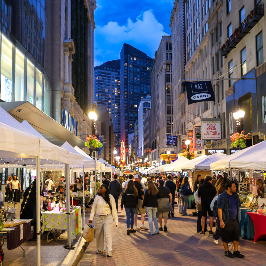 People shop at outdoor market stalls along a city street lined with tall buildings in the evening, under a blue sky with clouds. Lights illuminate the scene, and a crowd strolls between white tents.