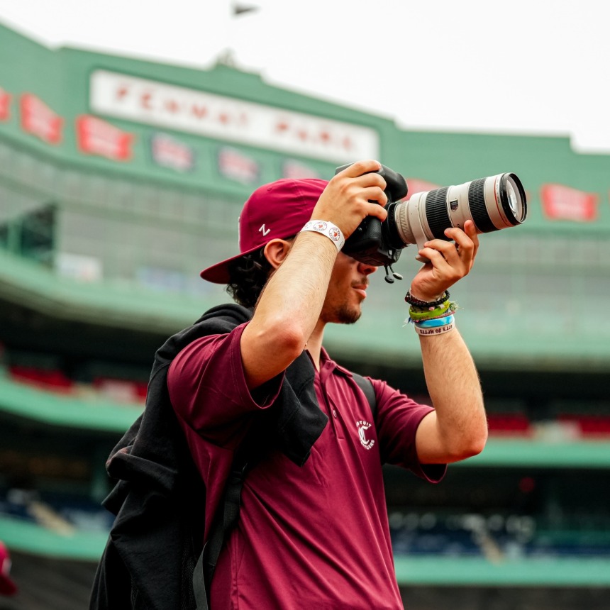 A person wearing a maroon shirt and cap takes a photo with a camera at Fenway Park. Stadium seating and the iconic Fenway Park sign are visible in the background.