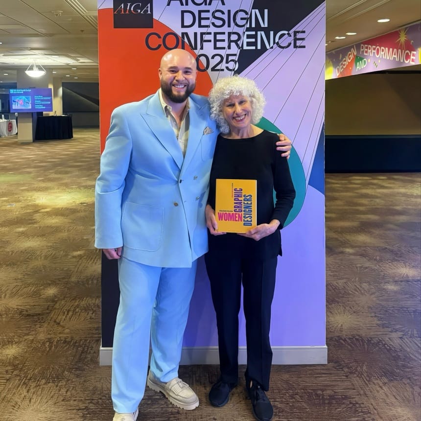 Two people stand and smile in front of an AIGA Design Conference 2025 sign. The person on the left wears a light blue suit; the person on the right holds a book titled “Women Graphic Designers.”.