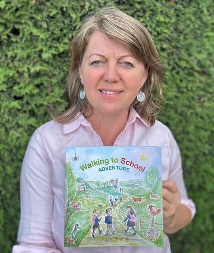 A woman with shoulder-length light brown hair, wearing a light pink shirt, stands outside in front of green foliage, holding up a children’s book titled Walking to School Adventure, ready to share her love of teaching and storytelling.