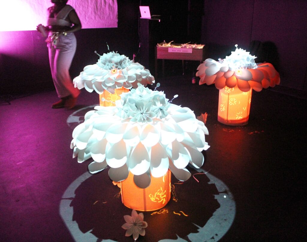 Three glowing sculptures resembling large, illuminated white flowers made of paper or plastic spoons are displayed in a dark room. A person stands in the background near a screen, partially lit by purple light.