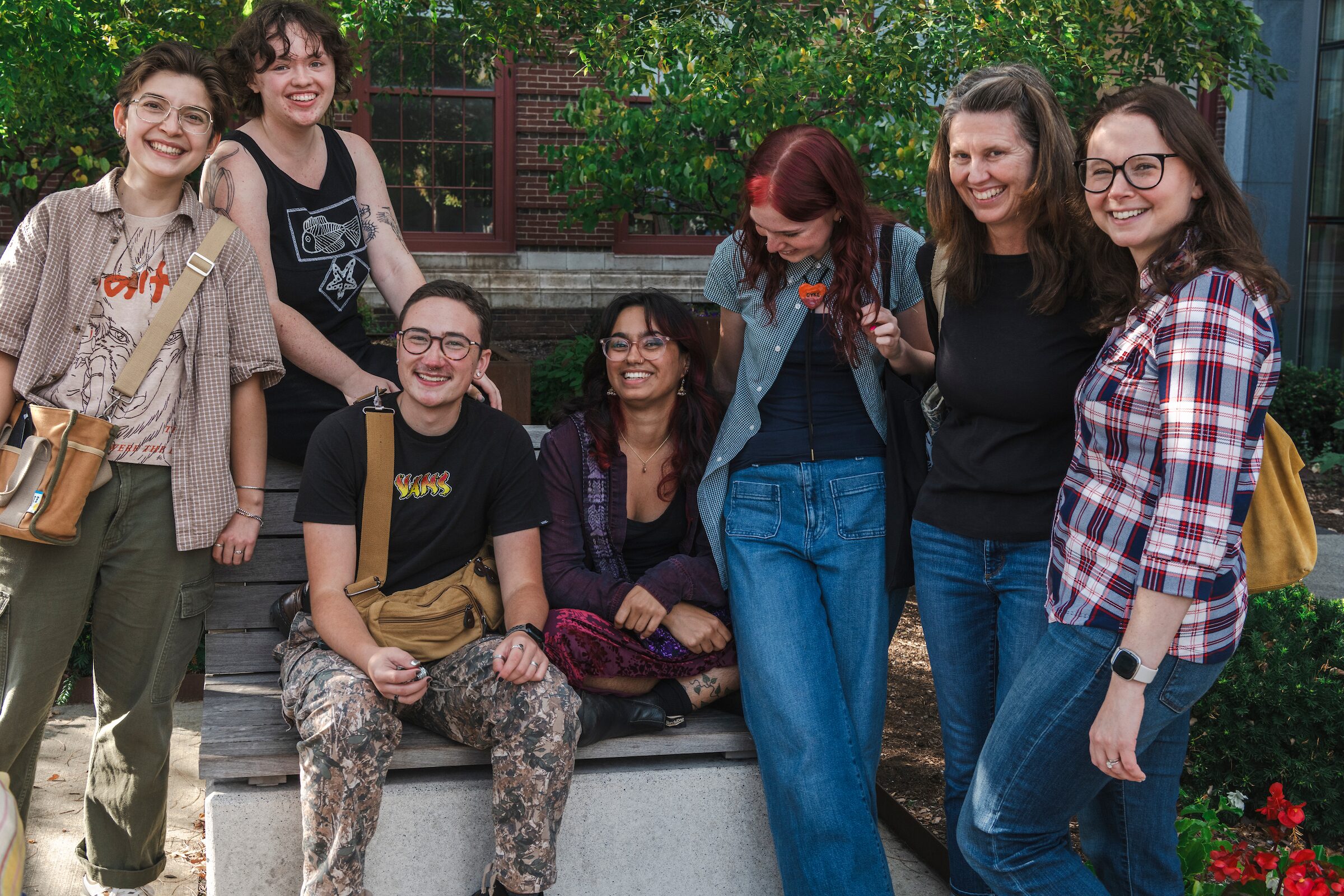A group of seven people smiling and posing together outdoors on a sunny day near a bench, surrounded by greenery and a brick building in the background—capturing a vibrant moment at MassArt.