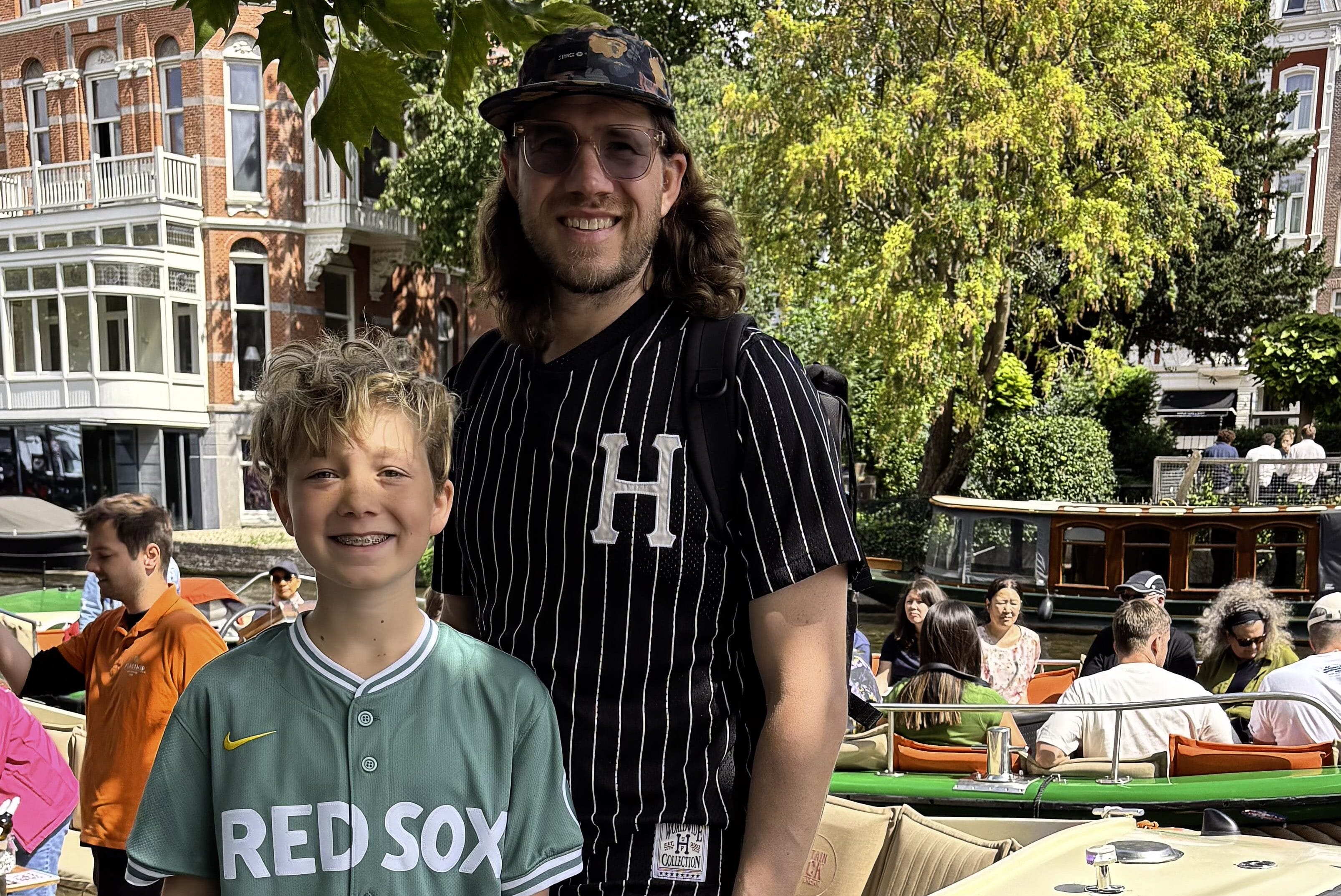 A smiling man and boy stand by a canal boat in a sunny city setting, enjoying their summer abroad. The boy wears a green Red Sox jersey and black shorts, while the man wears glasses, a black striped shirt, and a cap. People and trees fill the background.