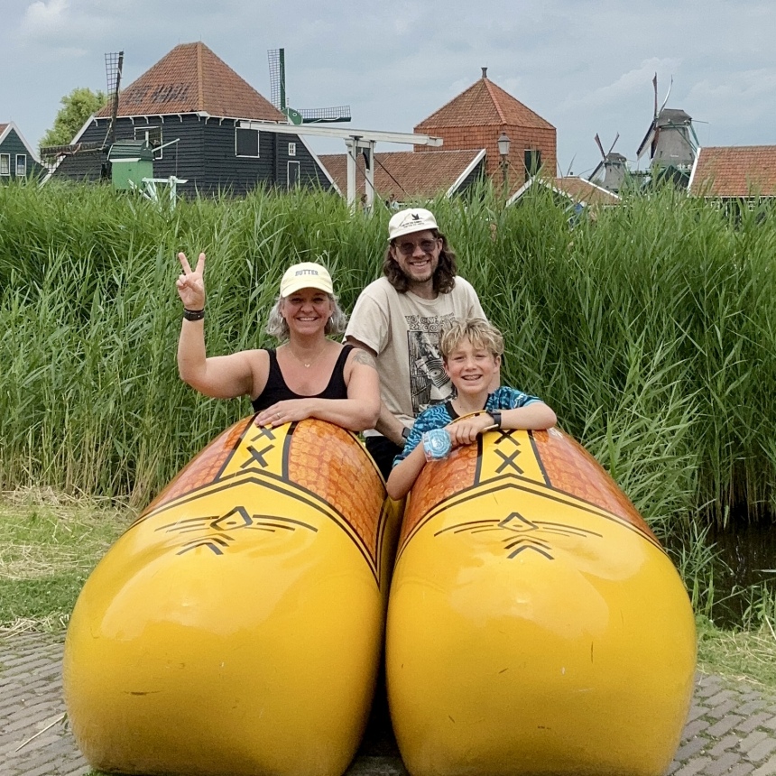 Three people, two adults and a child, smile inside giant yellow Dutch wooden shoe sculptures, with windmills and tall grass behind them—a perfect summer abroad memory. One adult flashes a peace sign as scrolls turning mark the start of their adventure.
