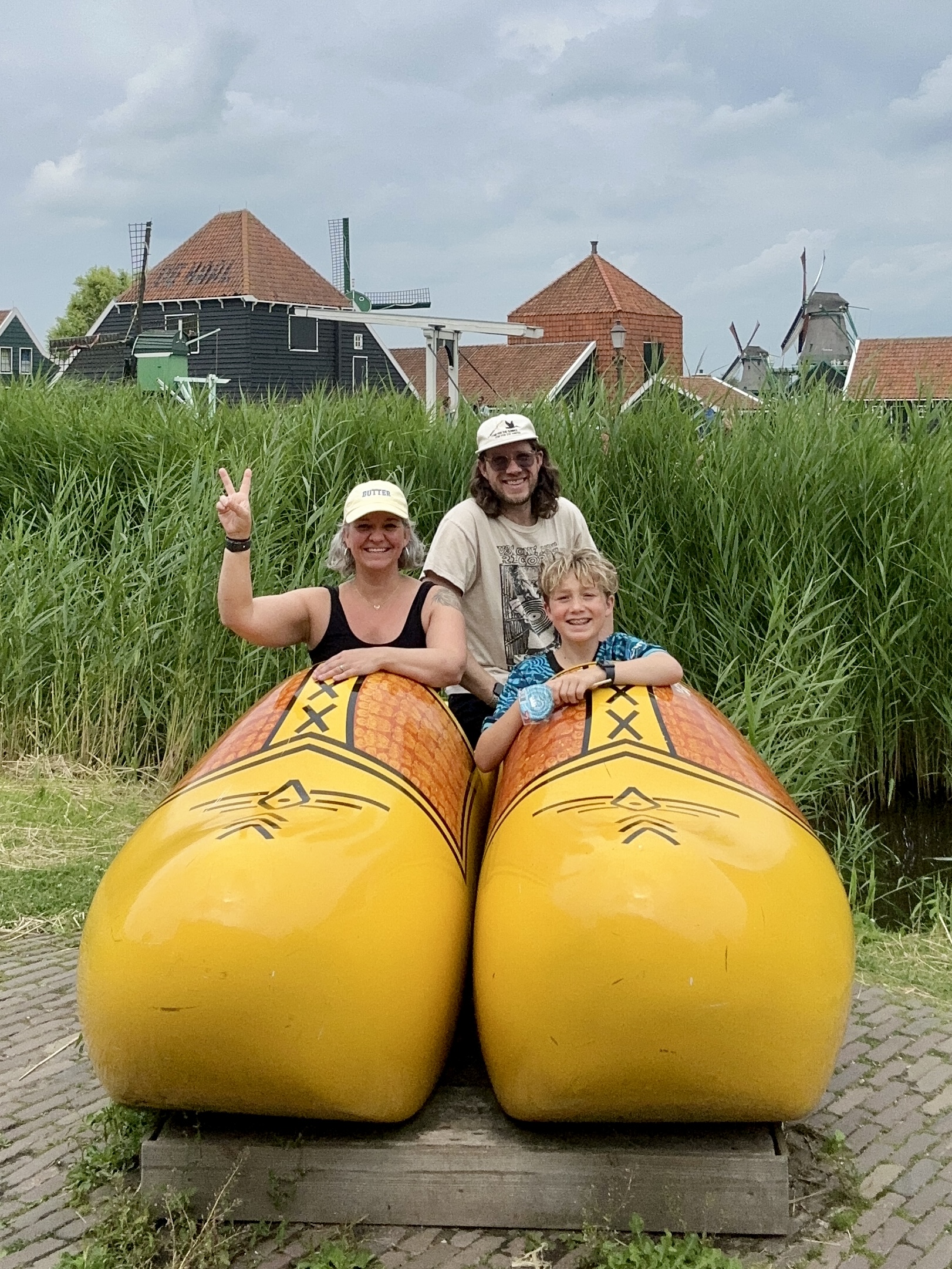 Three people, two adults and a child, smile inside giant yellow Dutch wooden shoe sculptures, with windmills and tall grass behind them—a perfect summer abroad memory. One adult flashes a peace sign as scrolls turning mark the start of their adventure.