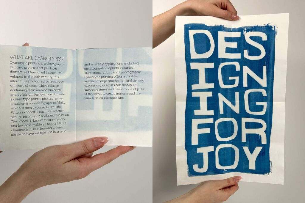 Two hands hold a folded paper. The left panel explains cyanotypes and their role in printmaking and art. The right panel features bold, white, hand-lettered text on blue that reads DESIGNING FOR JOY.