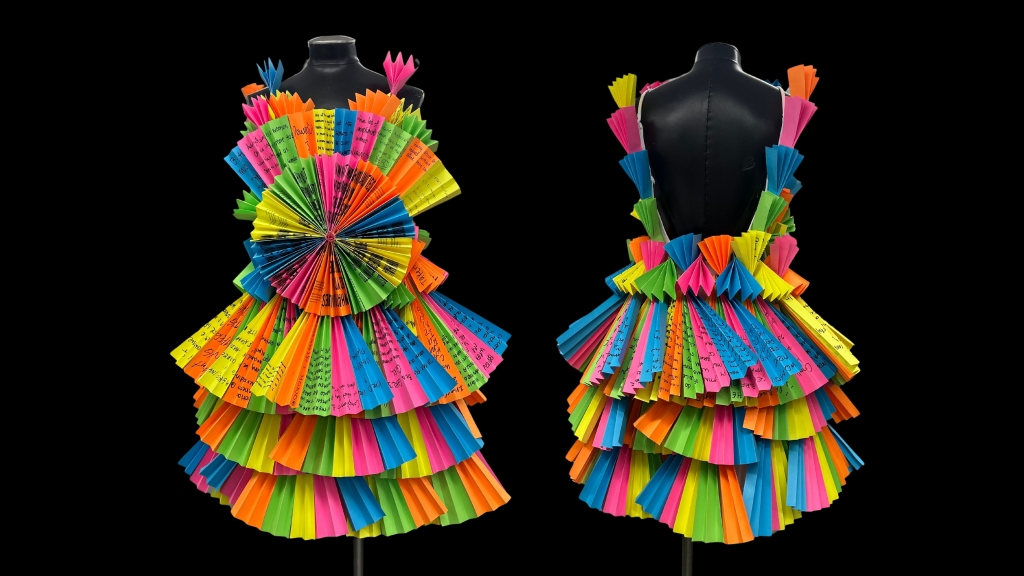 A colorful dress made of layered, pleated paper in neon orange, yellow, blue, green, and pink, displayed on a mannequin. The front features a large fan-shaped paper rosette at the waist. The background is black.