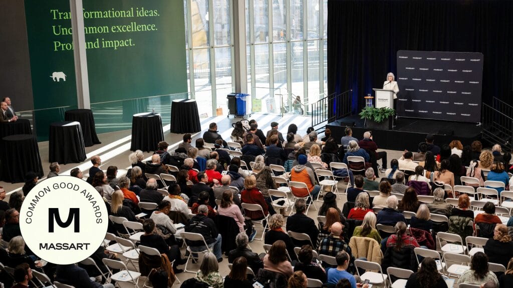A large audience sits in rows facing a stage where a speaker stands at a podium. Behind the speaker is a black backdrop with MassArt logos, while the MassArt Common Good Awards logo appears in the foreground. A green wall displays text about transformational ideas.