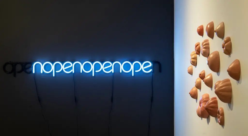 A neon sign on a dark wall in the MassArt Graduate Gallery displays “nopenopenopenope” in blue, with part of “open” visible. On a white wall nearby, sculpted flesh-toned human ears cluster—a glimpse into innovative exhibition strategies.