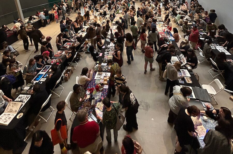 A crowded indoor convention where lively meetings and events unfold, with many people browsing and selling items at long rows of tables covered with art, books, and merchandise. The scene buzzes as attendees walk, chat, and enjoy the vibrant admissions experience.