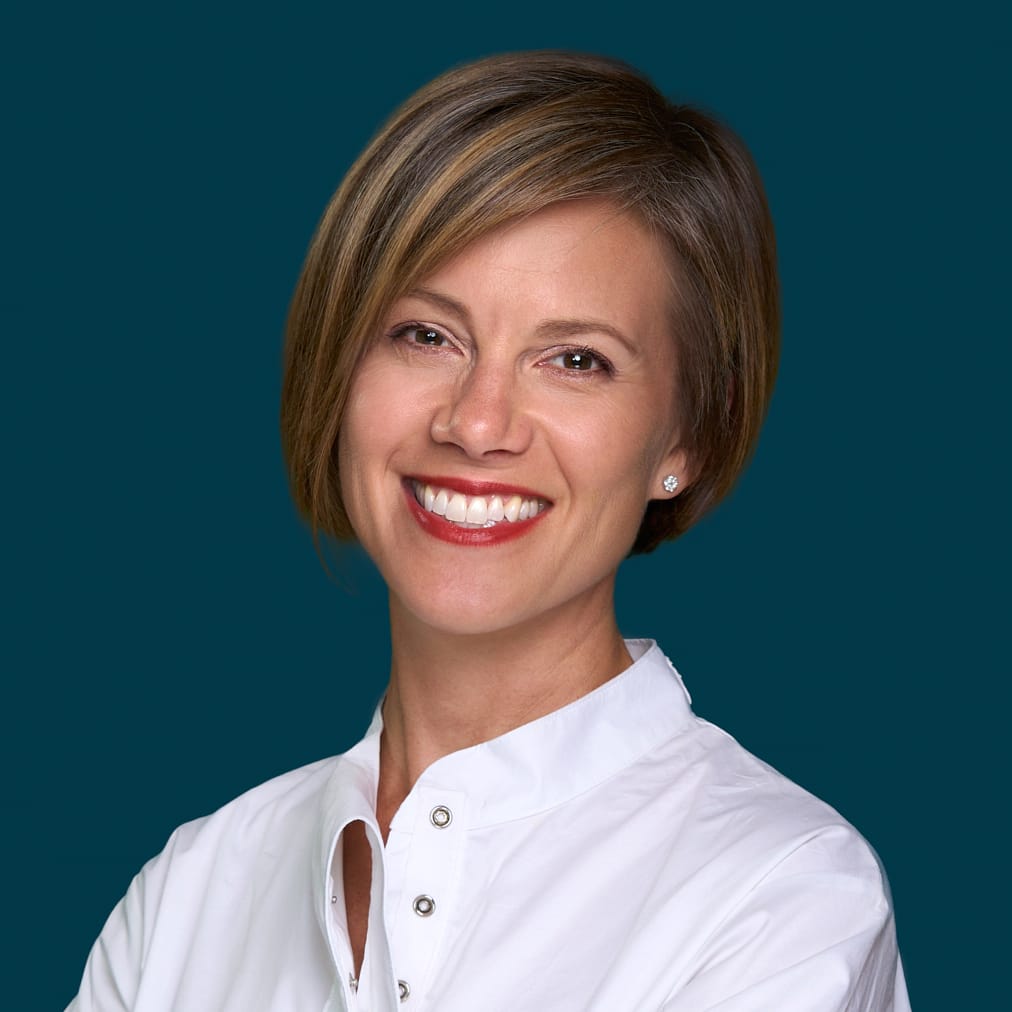 A woman with short brown hair smiles warmly, wearing a white buttoned shirt and stud earrings, standing against a solid teal background, representing MassArt at the Common Good Awards.