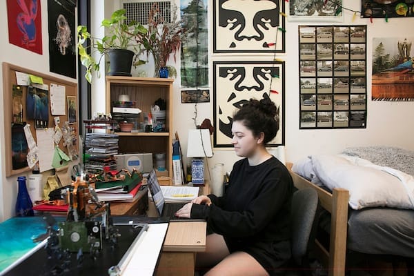 A person sits at a desk typing on a laptop in a cozy, decorated dorm room with posters, plants, and books—perhaps preparing for upcoming Admissions Meetings. The bed is to the right, and natural light comes from a window on the left.