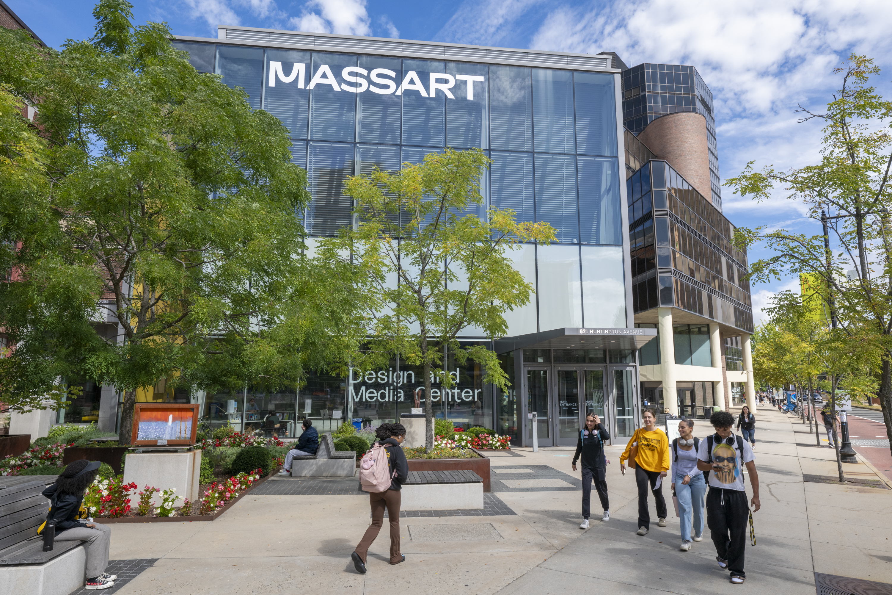 Several people walk in front of the MassArt Design and Media Center, reflecting why MassArt is considered one of the top art schools. The modern glass building is surrounded by trees, flowers, and benches under a blue sky with clouds.