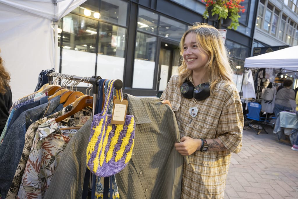 A smiling young woman in a plaid shirt stands at a MassArt Night Market booth in Downtown Crossing, surrounded by racks of clothing and a colorful knit bag. She wears headphones around her neck, showcasing local art & design talent among shops and tents in the background.