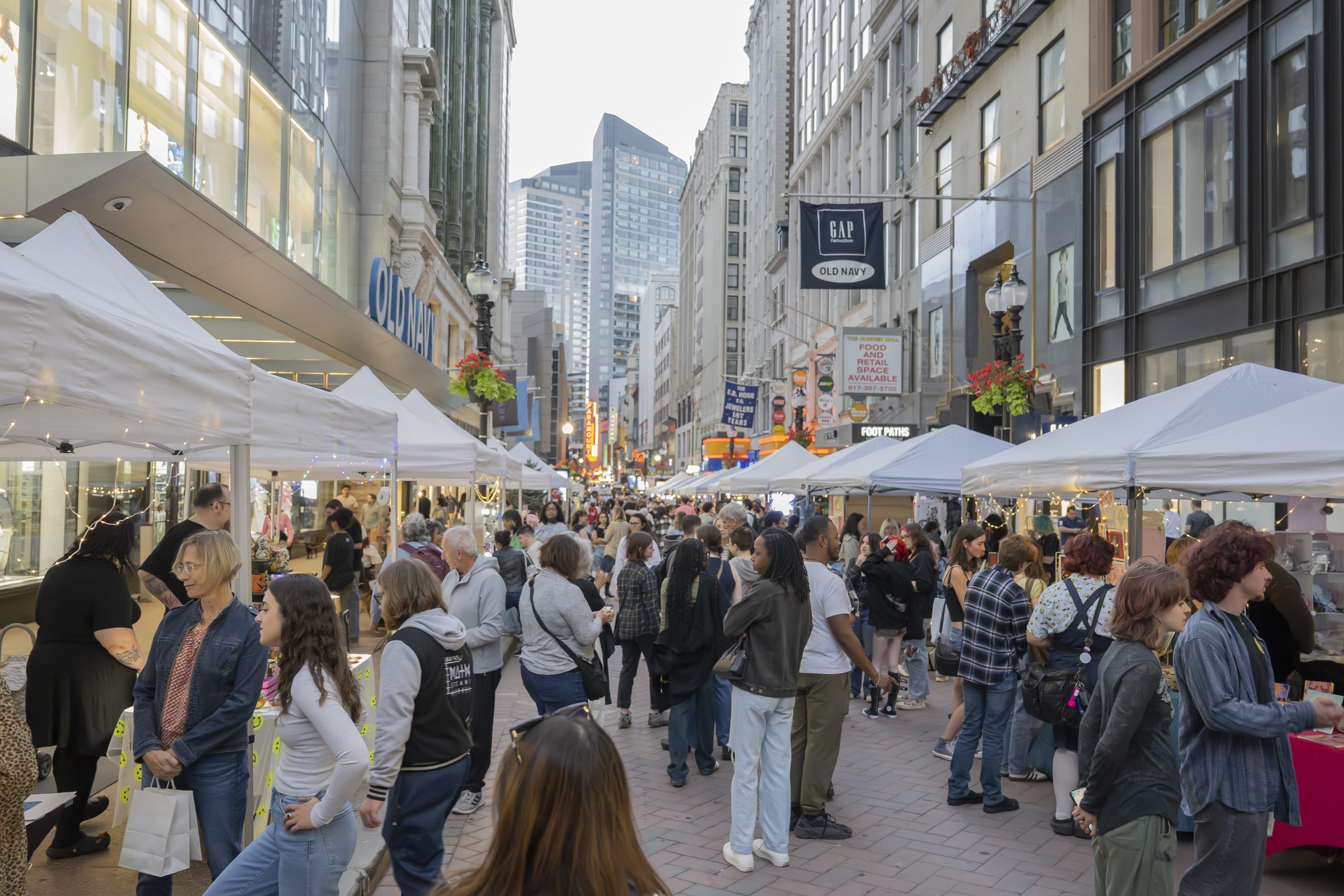 A busy street market with white tents lined on both sides, crowded with people shopping and browsing at the vibrant Downtown Crossing, with city storefronts and Art & Design Talent from MassArt Night Market on display.