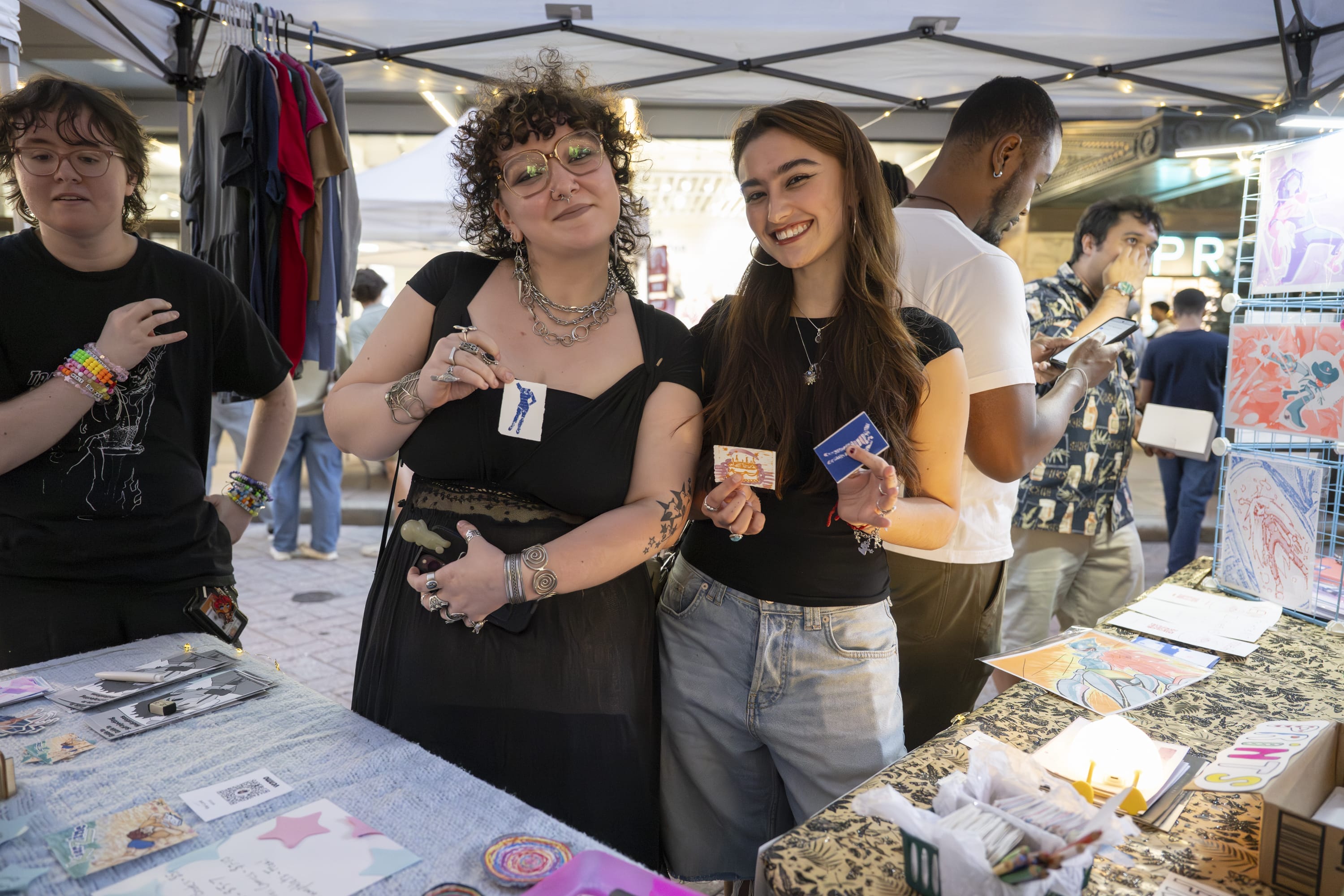 Two people smile and pose at a MassArt Night Market stall in Downtown Crossing, holding cards or stickers. The table before them displays colorful art and crafts, showcasing local Art & Design Talent as others browse under a canopy in the background.