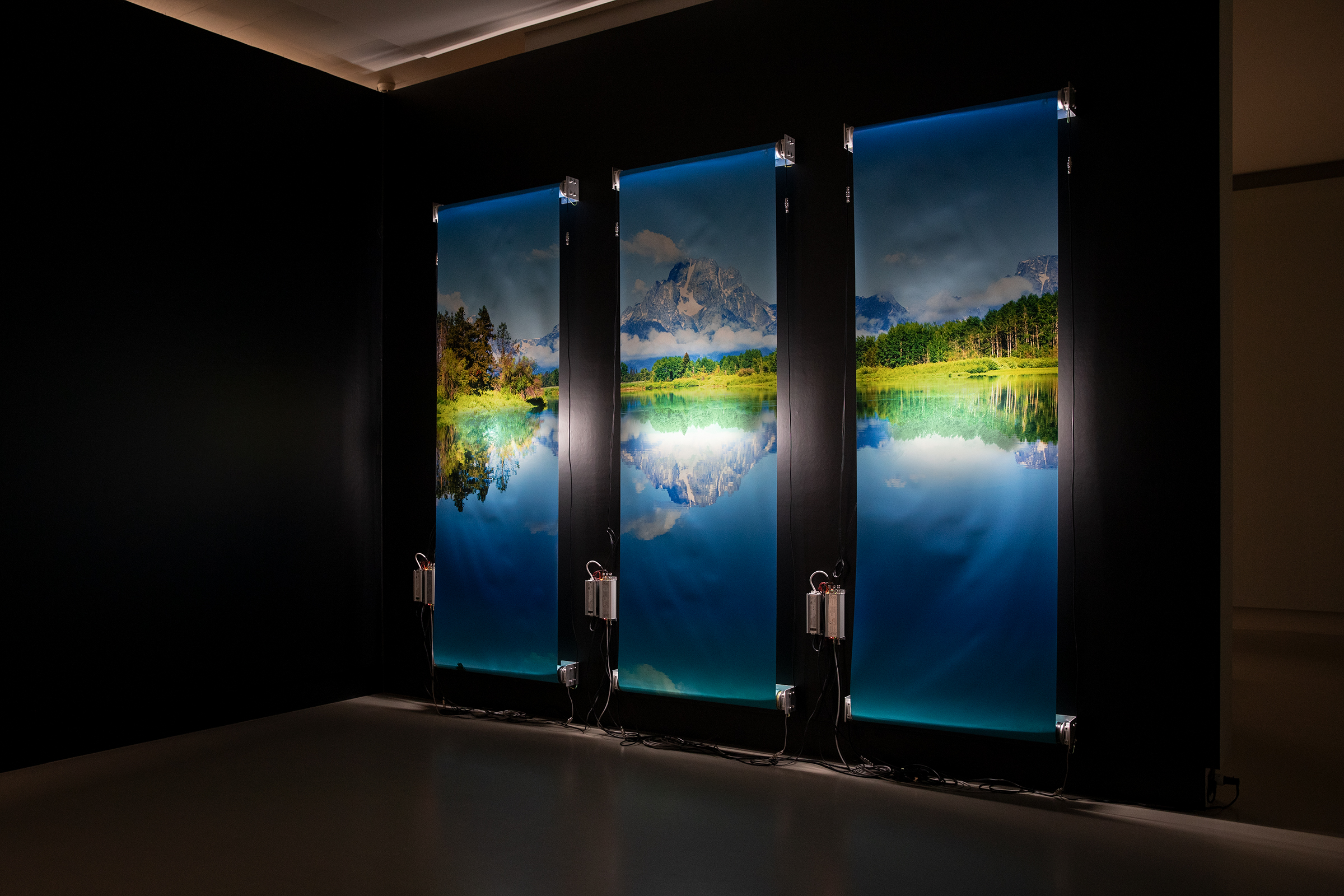 Three vertically-oriented illuminated panels display a serene landscape of mountains, trees, and a lake with reflections, evoking memories of a summer abroad. Set against a dark gallery wall, electrical equipment is visible beneath each panel.