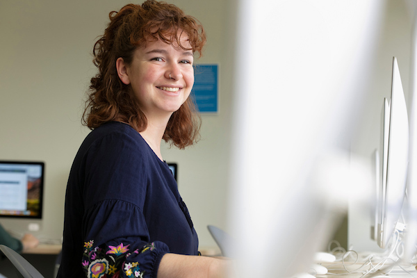 A young woman with curly brown hair and a dark patterned blouse smiles while sitting at a desk with computer monitors, preparing for Admissions Meetings in a bright, modern classroom or computer lab.