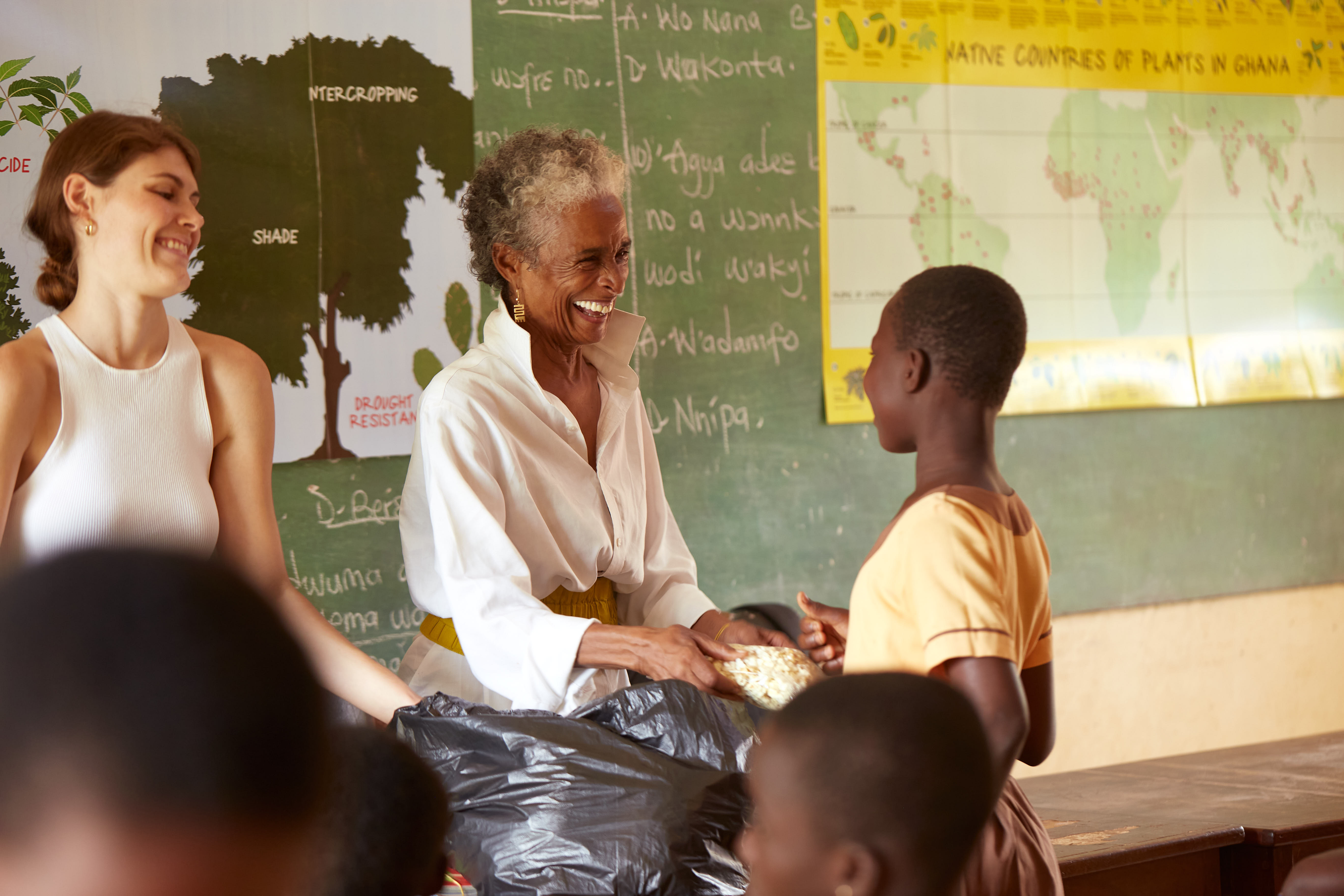 A smiling older woman hands a small bag of food to a child in a classroom, while another woman looks on. Children are in the foreground, and educational posters promote the Tyrone Maurice Adderley Lecture Series on the walls behind them.