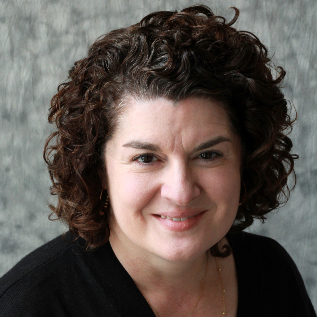 A woman with short, curly dark brown hair smiles gently at the camera. She is wearing a black top and a subtle gold necklace, with a soft gray, textured background—capturing the spirit celebrated by MassArt’s Common Good Awards.