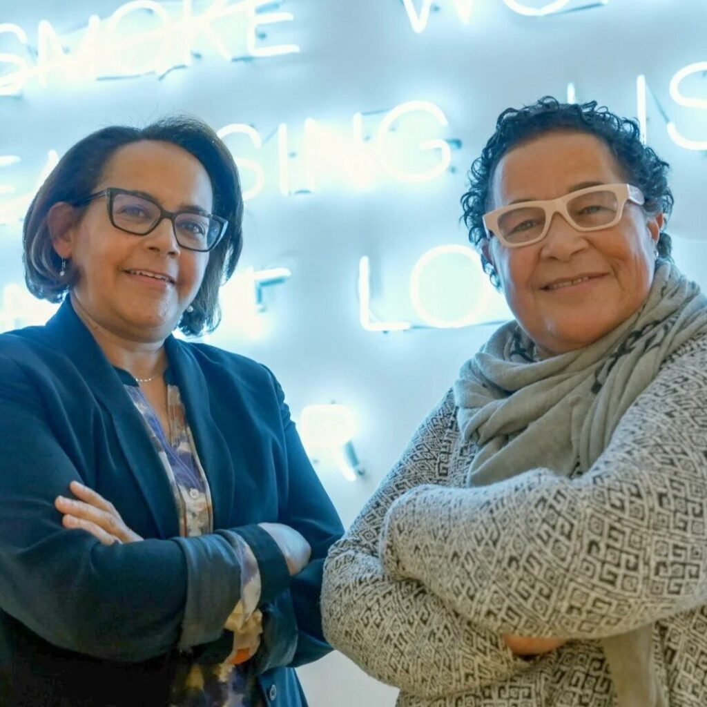 Two women smiling with arms crossed stand before a bright blue neon sign at MassArt. Both wear glasses and casual clothing—one in a scarf and patterned sweater, the other in a blazer—celebrating the spirit of the Common Good Awards.