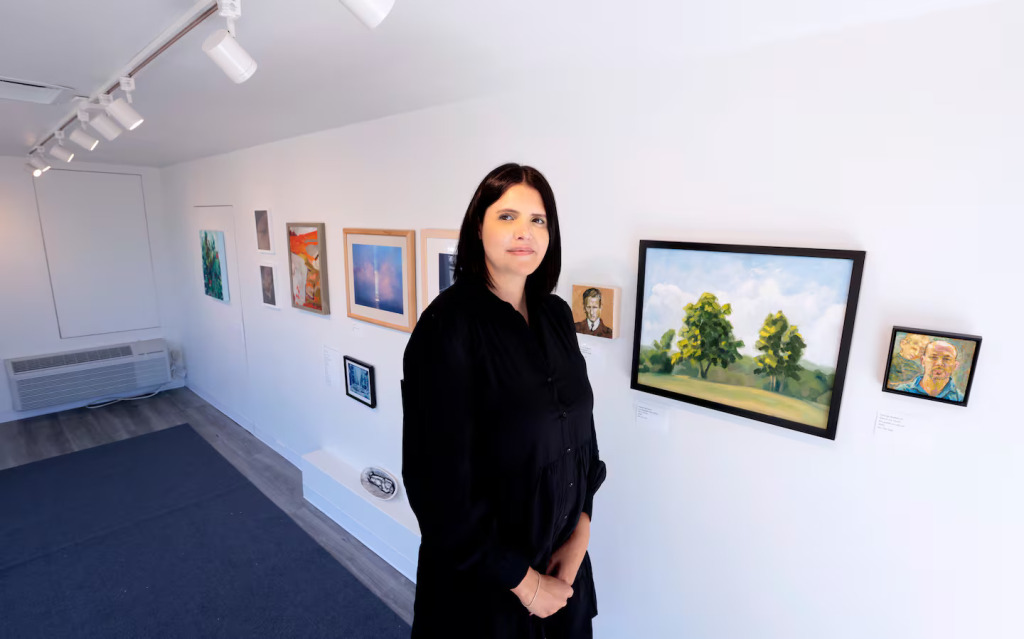 A woman in a black dress stands in a brightly lit art gallery, surrounded by paintings and artwork created by local artists displayed on white walls.